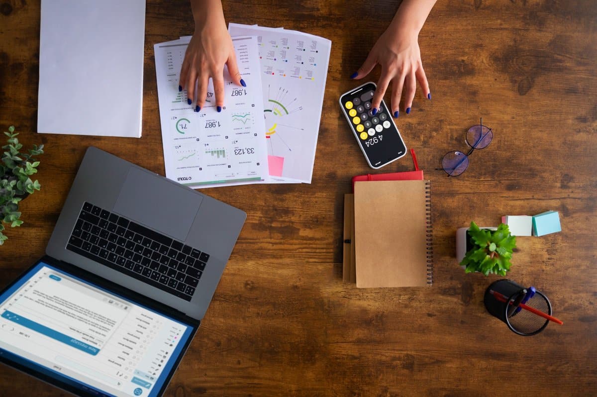 Top-view workspace showing a person organizing personal finances, analyzing printed financial charts, using a calculator app on a smartphone, and working with a laptop, notebooks, glasses, sticky notes, and a small plant on a wooden desk.