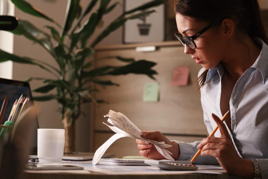 Woman sitting at a desk reviewing a stack of receipts while using a calculator, with office supplies, notes, and a soft-lit home office environment around her, symbolizing the importance of understanding your financial situation by closely tracking expenses and analyzing monthly spending habits.