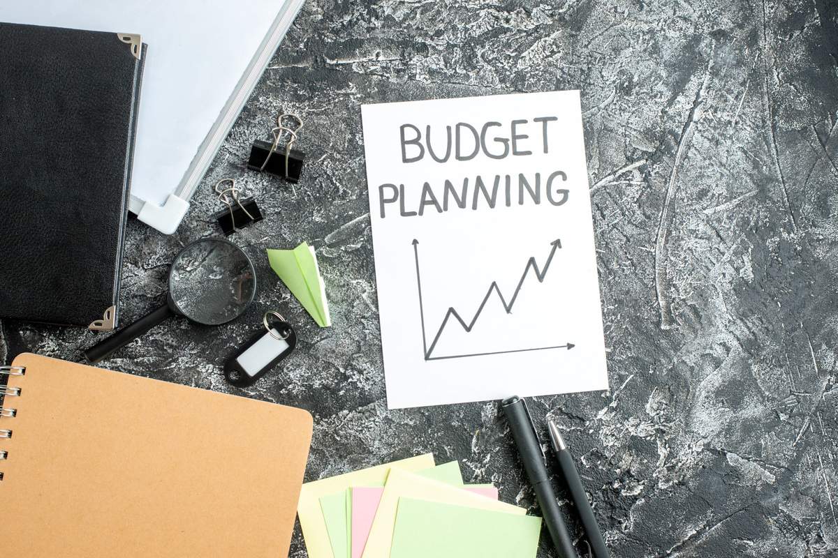 Flat-lay scene featuring a ‘Budget Planning’ sheet with an upward trending graph, surrounded by notebooks, pens, binder clips, sticky notes, and a magnifying glass on a textured gray surface, representing how to create an efficient monthly budget through organized planning and clear financial structuring.